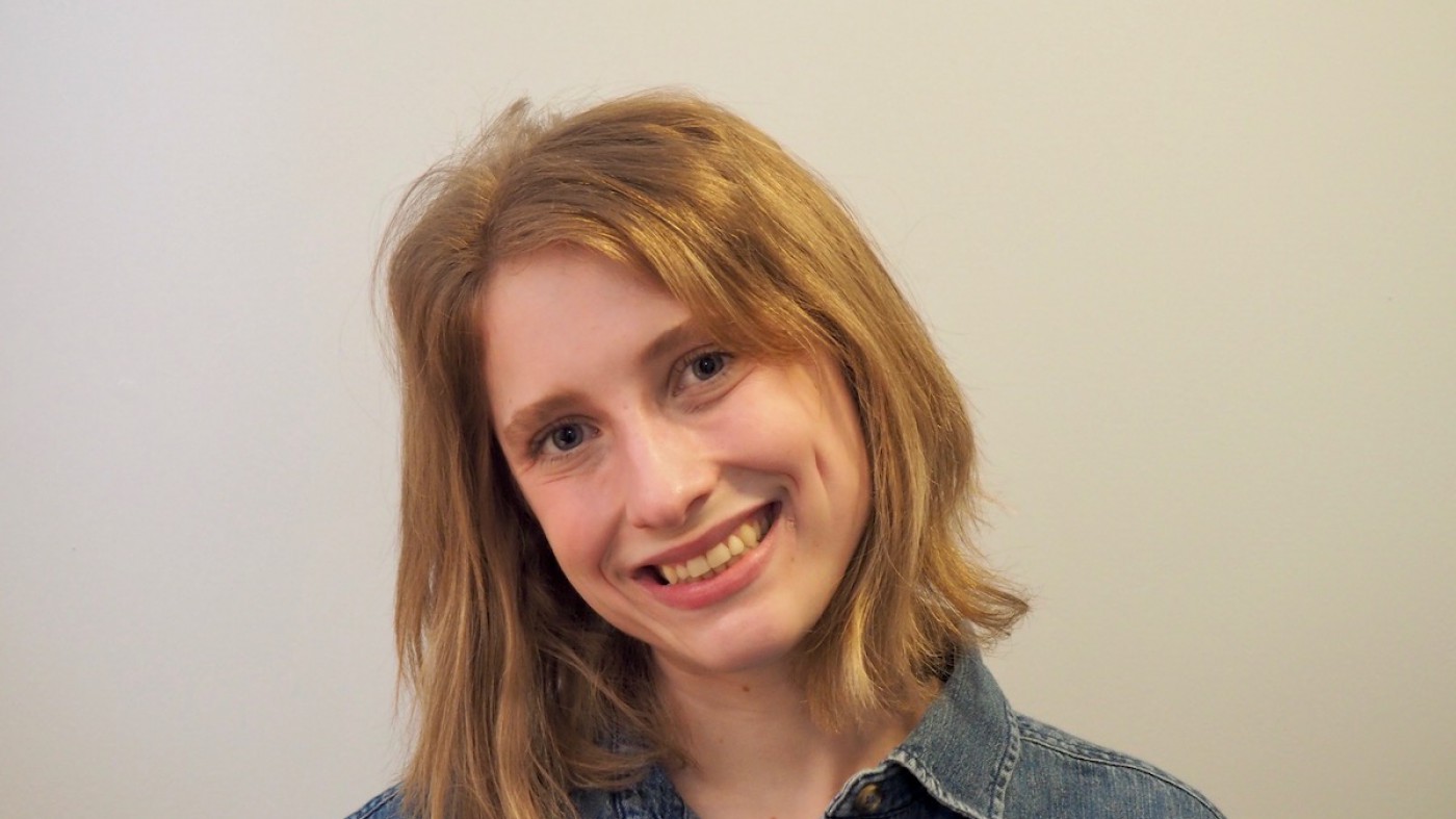 A photo of a person smiling and tilting their head to the side. They are wearing a jean shirt and have shoulder-length blond hair.