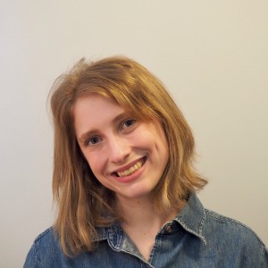 A photo of a person smiling and tilting their head to the side. They are wearing a jean shirt and have shoulder-length blond hair.