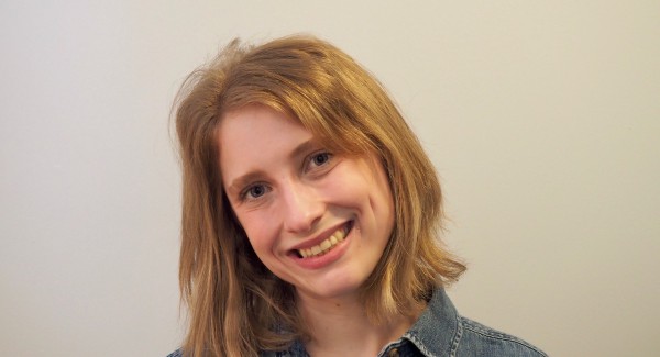 A photo of a person smiling and tilting their head to the side. They are wearing a jean shirt and have shoulder-length blond hair.