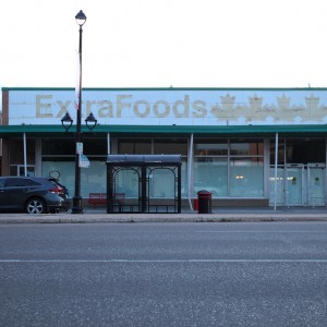 An exterior shot of the now-closed Extra Foods grocery store on Broadway Avenue in Saskatoon. The view from across a paved street is of an off-white building with a faded sign across the front that says: Extra Foods, accompanied by a graphic of four maple leaves.