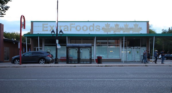 An exterior shot of the now-closed Extra Foods grocery store on Broadway Avenue in Saskatoon. The view from across a paved street is of an off-white building with a faded sign across the front that says: Extra Foods, accompanied by a graphic of four maple leaves.