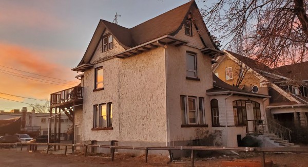 A photo of a white stucco house with a brown roof, next to an empty lot. Behind it, the sun is setting and the sky is pink and orange.