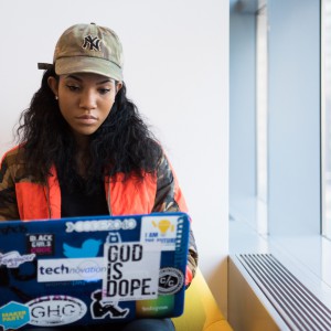 A woman sits on a couch working on an open laptop. She is wearing a tan New York Yankees hat, has long wavy black hair, and is wearing an orange and camouflage patterned jacket.