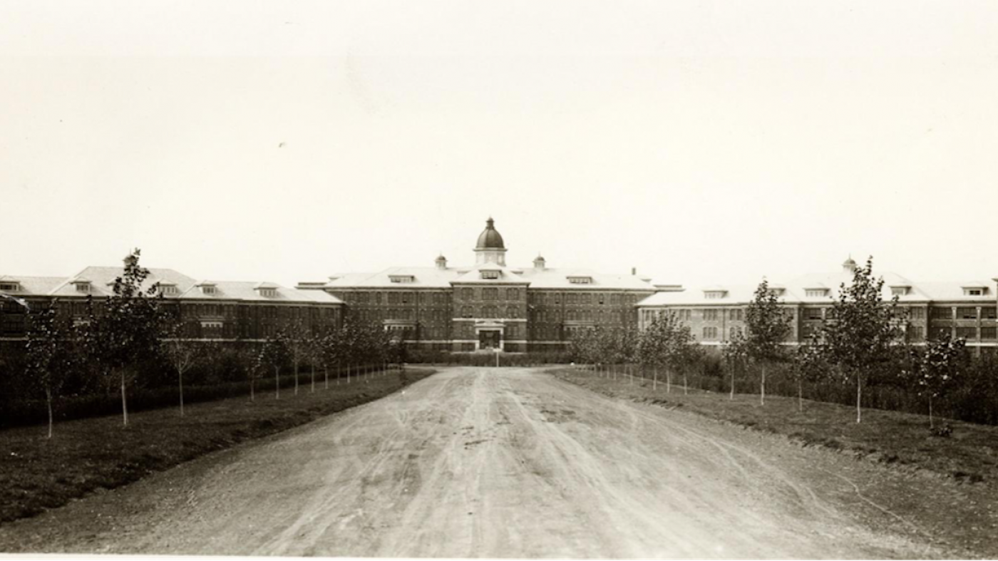 An old black-and-white photo of the Saskatchewan Provincial Mental Hospital in Weyburn. There is a long, broad driveway leading up to a large brick building, with a few trees lining the road.