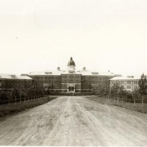 An old black-and-white photo of the Saskatchewan Provincial Mental Hospital in Weyburn. There is a long, broad driveway leading up to a large brick building, with a few trees lining the road.