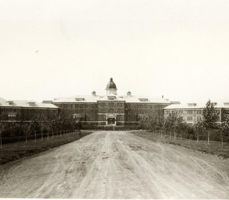 An old black-and-white photo of the Saskatchewan Provincial Mental Hospital in Weyburn. There is a long, broad driveway leading up to a large brick building, with a few trees lining the road.