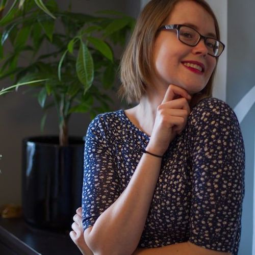 A photo of Shelby Lyn Lowe. Shelby has a straight brown hair in a bob, and glasses. She is wearing a navy shirt, red lipstick, and glasses. She is resting her hand on her chin and smiling sideways at the camera.