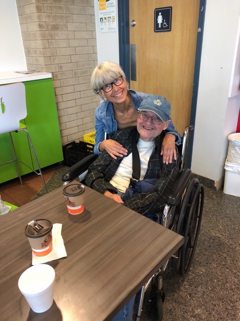 Two people are pictured beside each other, both smiling. The person on the right is sitting in a wheelchair.